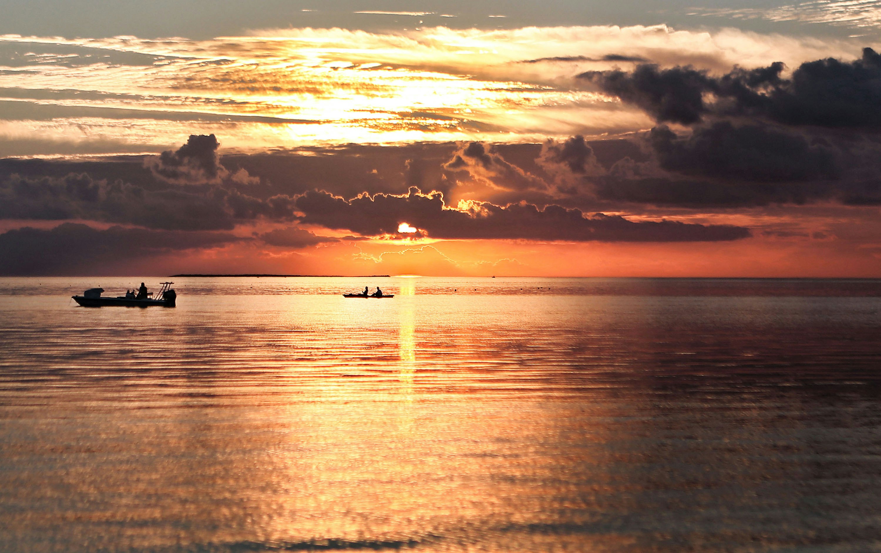 Sunset over calm ocean with boats silhouetted against an orange and gold sky, emphasizing serene water reflections