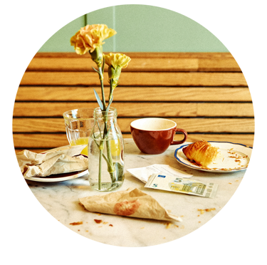 Frukostbord med blommor i vas, en kopp, croissant på tallrik och en fem-eurosedel.