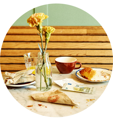Blommor i vas bredvid kaffekopp, croissant och eurosedel på cafébord.