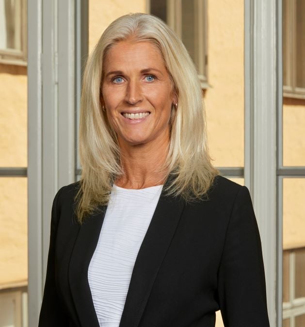 En kvinna med blont hår i en svart kavaj står framför ett fönster.