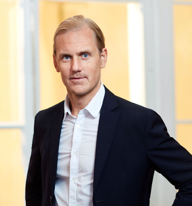 Man i svart kavaj och vit skjorta står framför ljusa fönster.