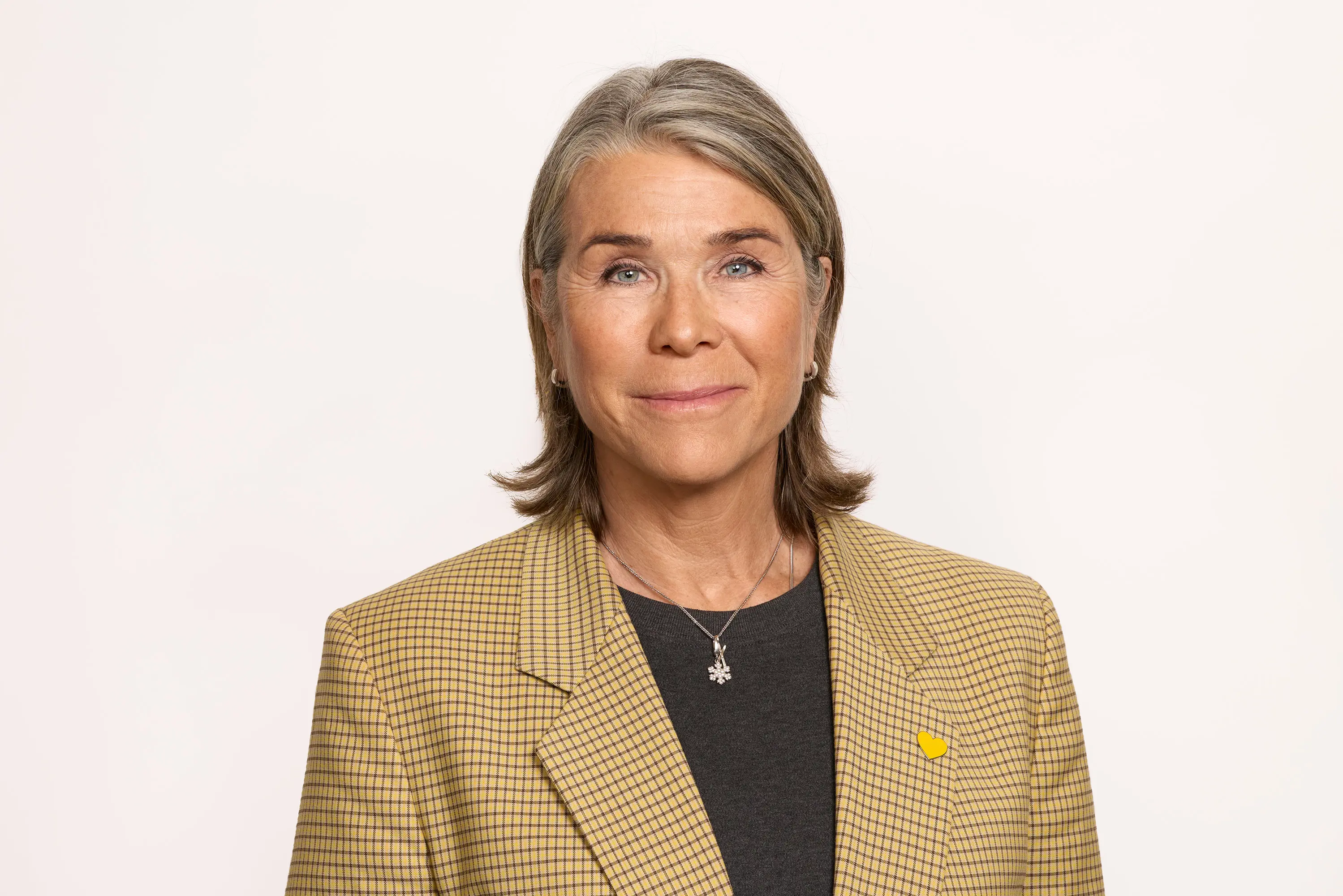 Professional woman in a plaid blazer posing against a plain background, wearing a necklace and a small yellow heart pin.