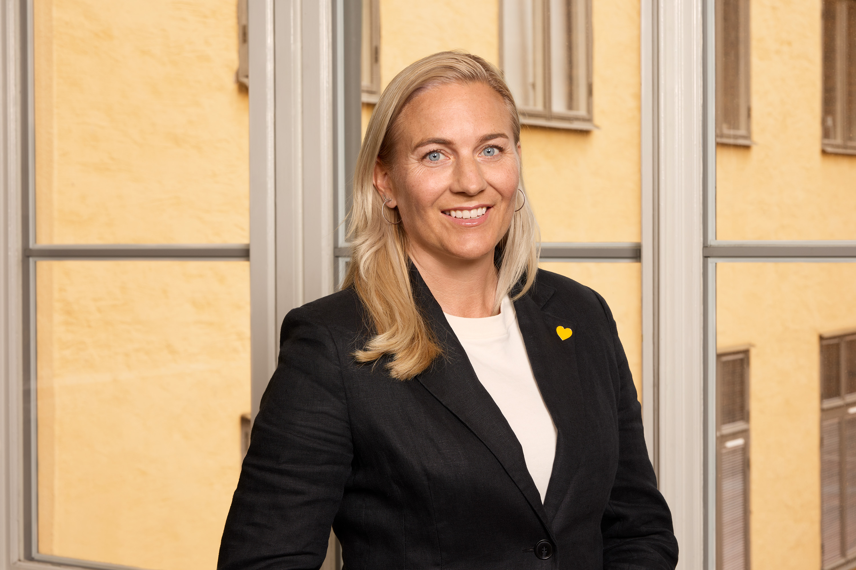 Kvinna i affärskostym med gul nål står i ett rum med stora fönster och gula väggar.