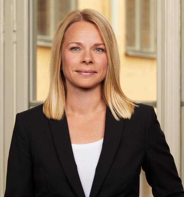 Kvinna i svart kavaj med långt blont hår står framför ett fönster.