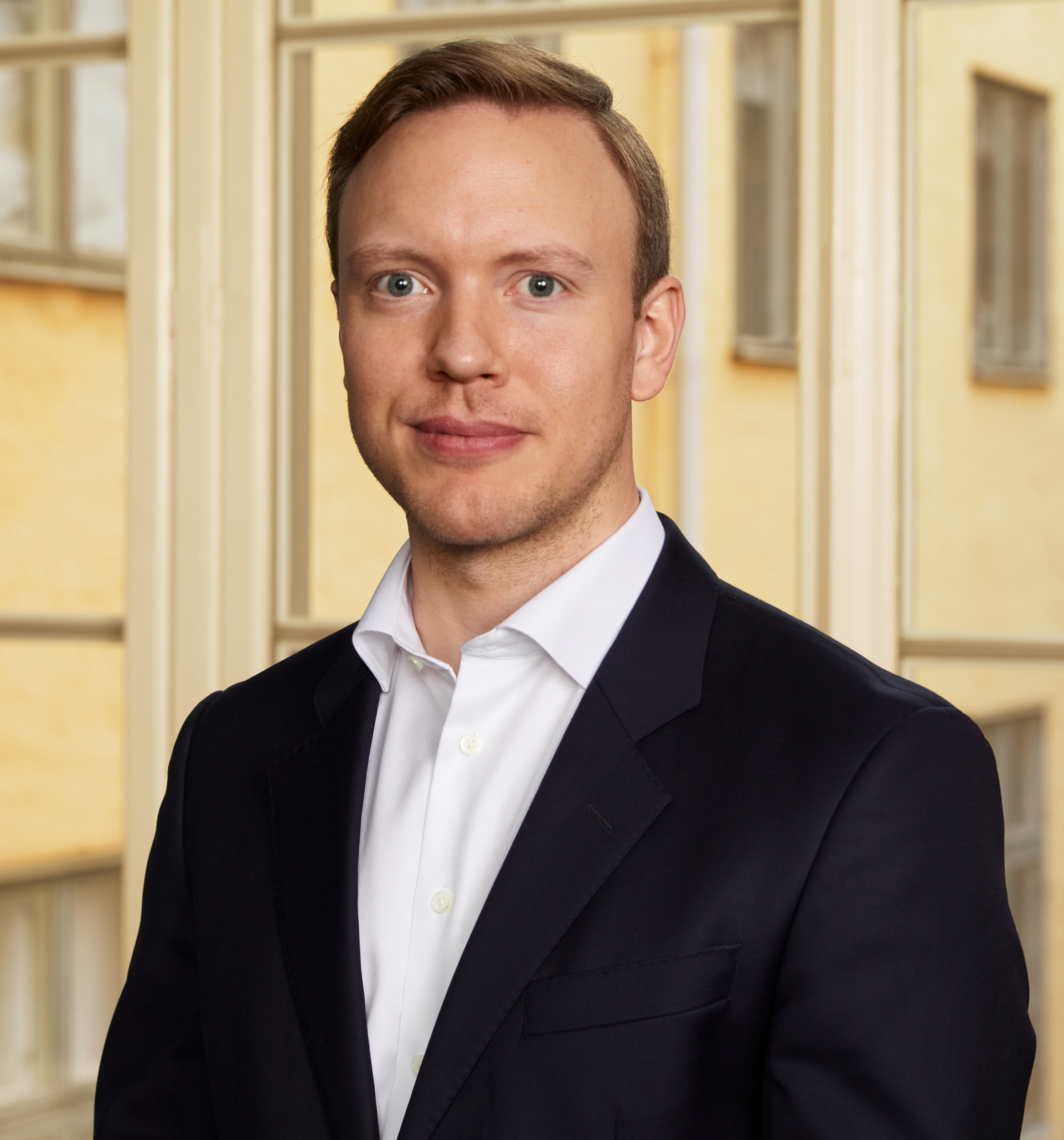 Person i kostym med kort blondt hår framför en suddig bakgrund av byggnader.