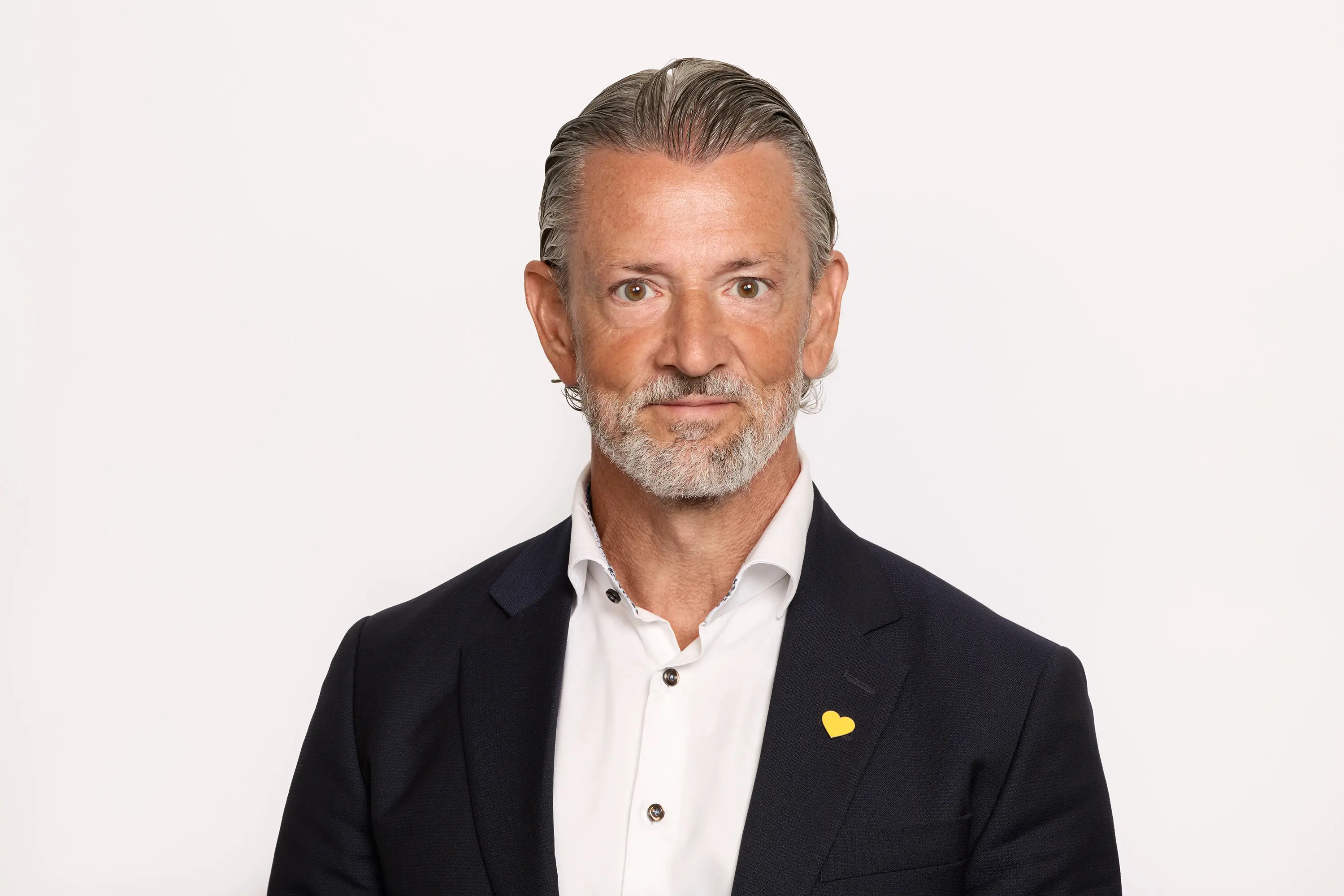 Man i kostym med vit skjorta och gul knappnål, porträtt mot vit bakgrund.