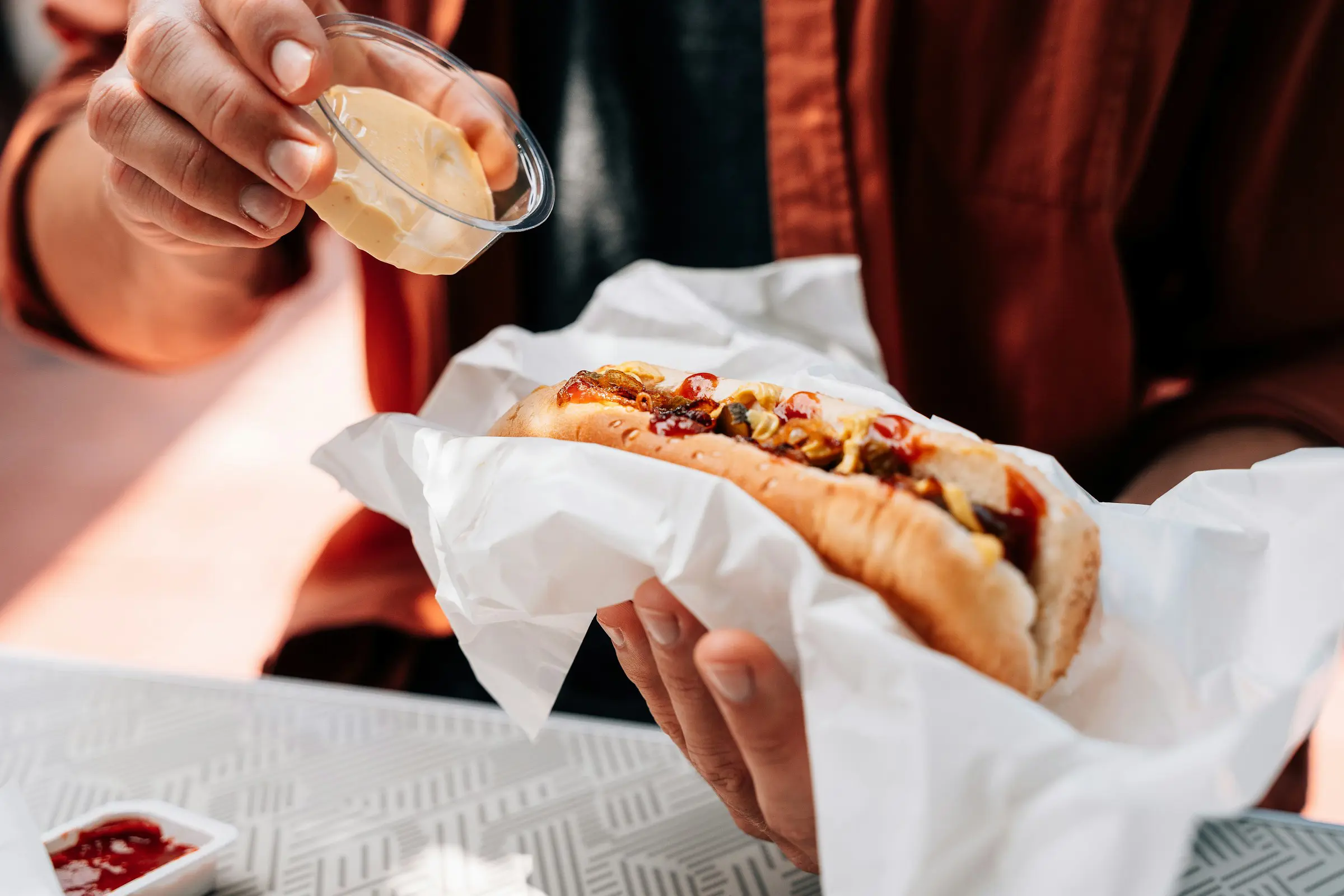 Person lägger på sås på en korv med bröd, garnerad med lök och ketchup, insvept i papper.