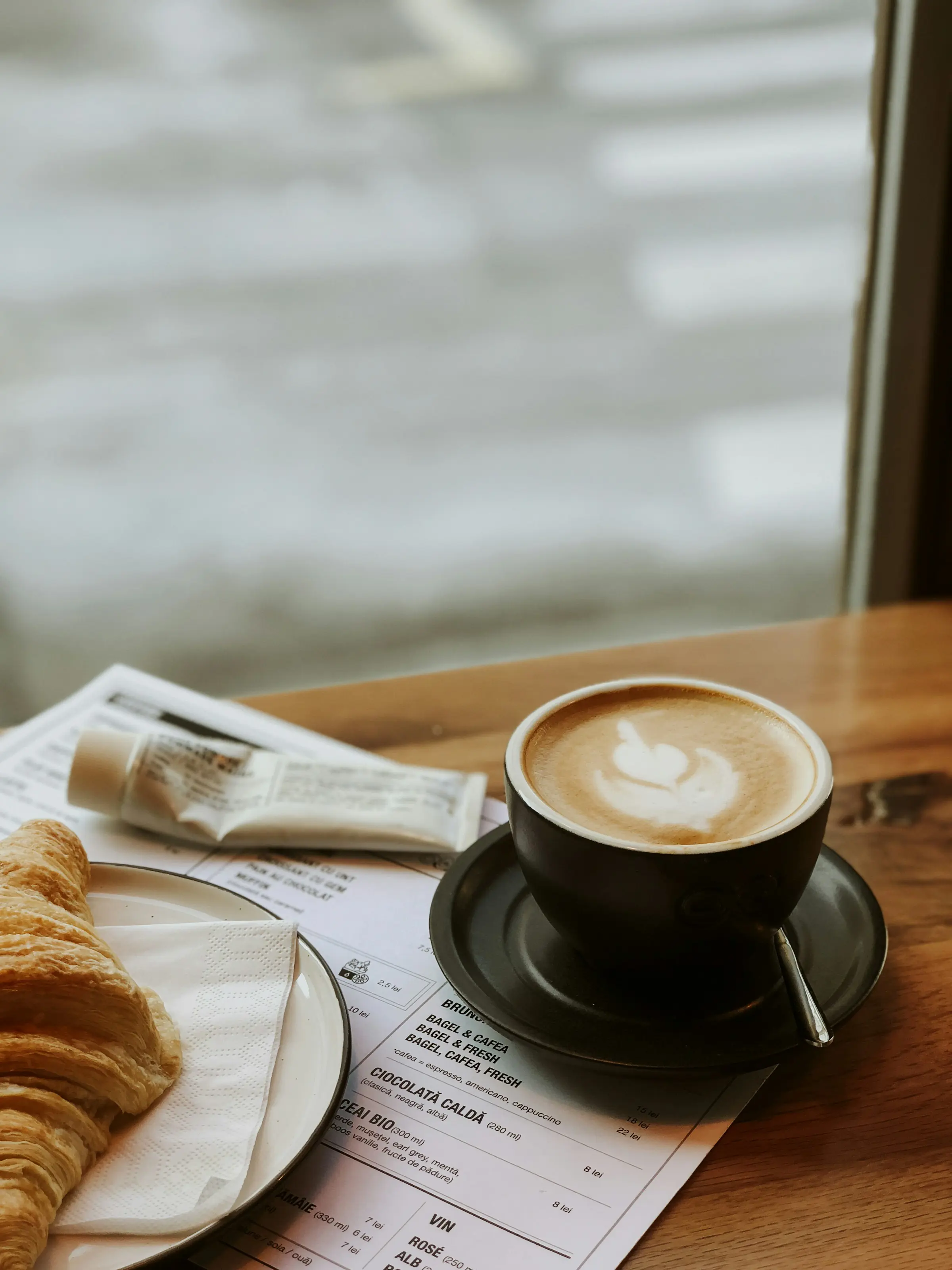 Cappuccino och croissant på träbord med meny i bakgrunden. Perfekt för frukost eller fika i café.