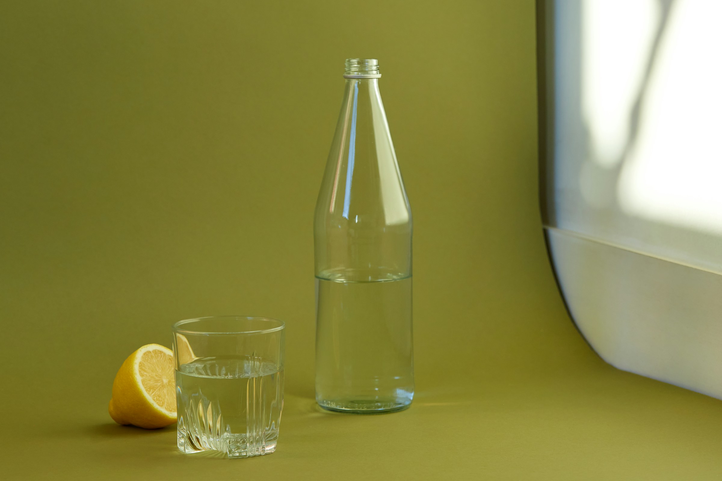 Glass bottle and tumbler of water with a lemon half on an olive green background.