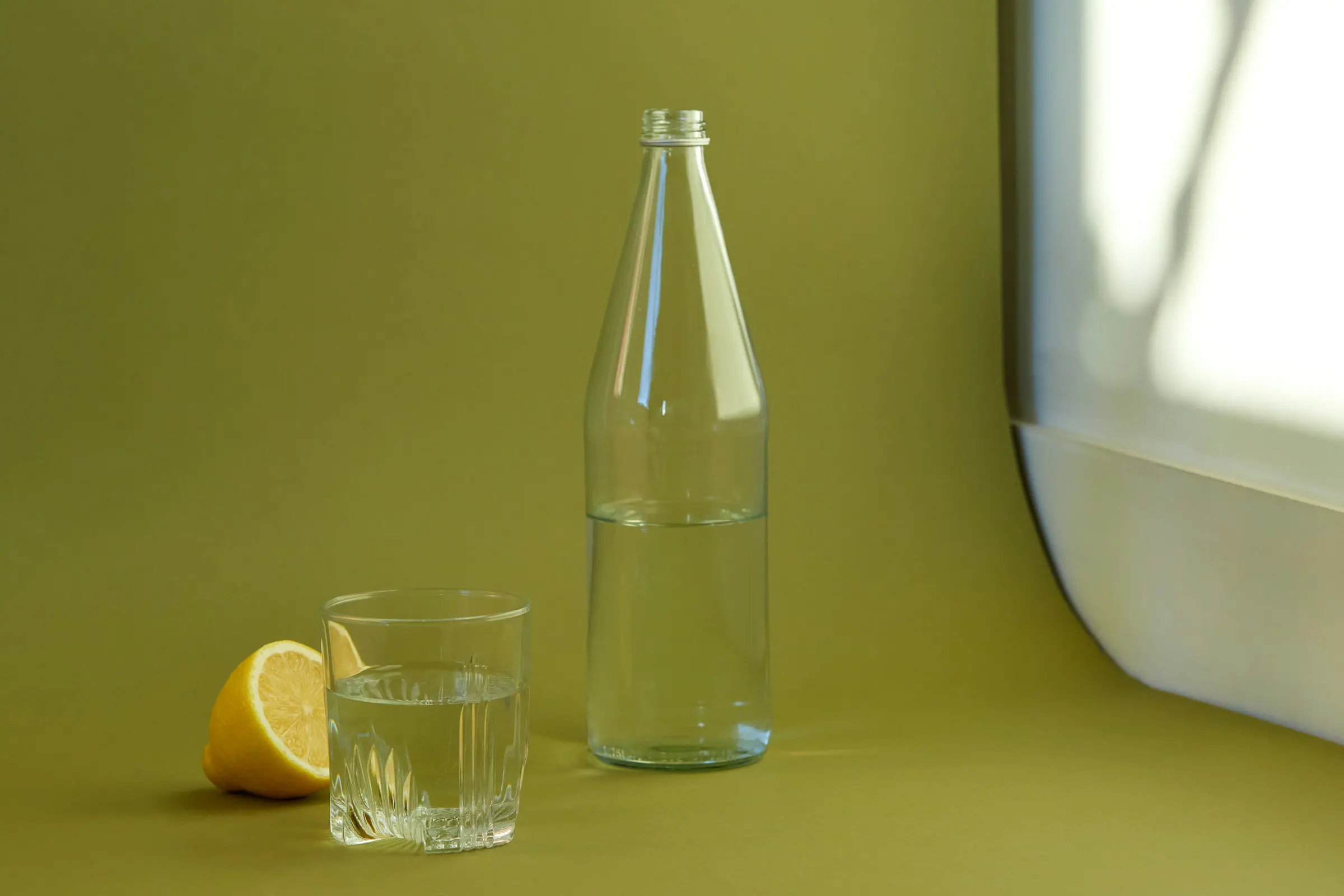 Glasflaska och glas med vatten bredvid en halverad citron på ett grönt bord.