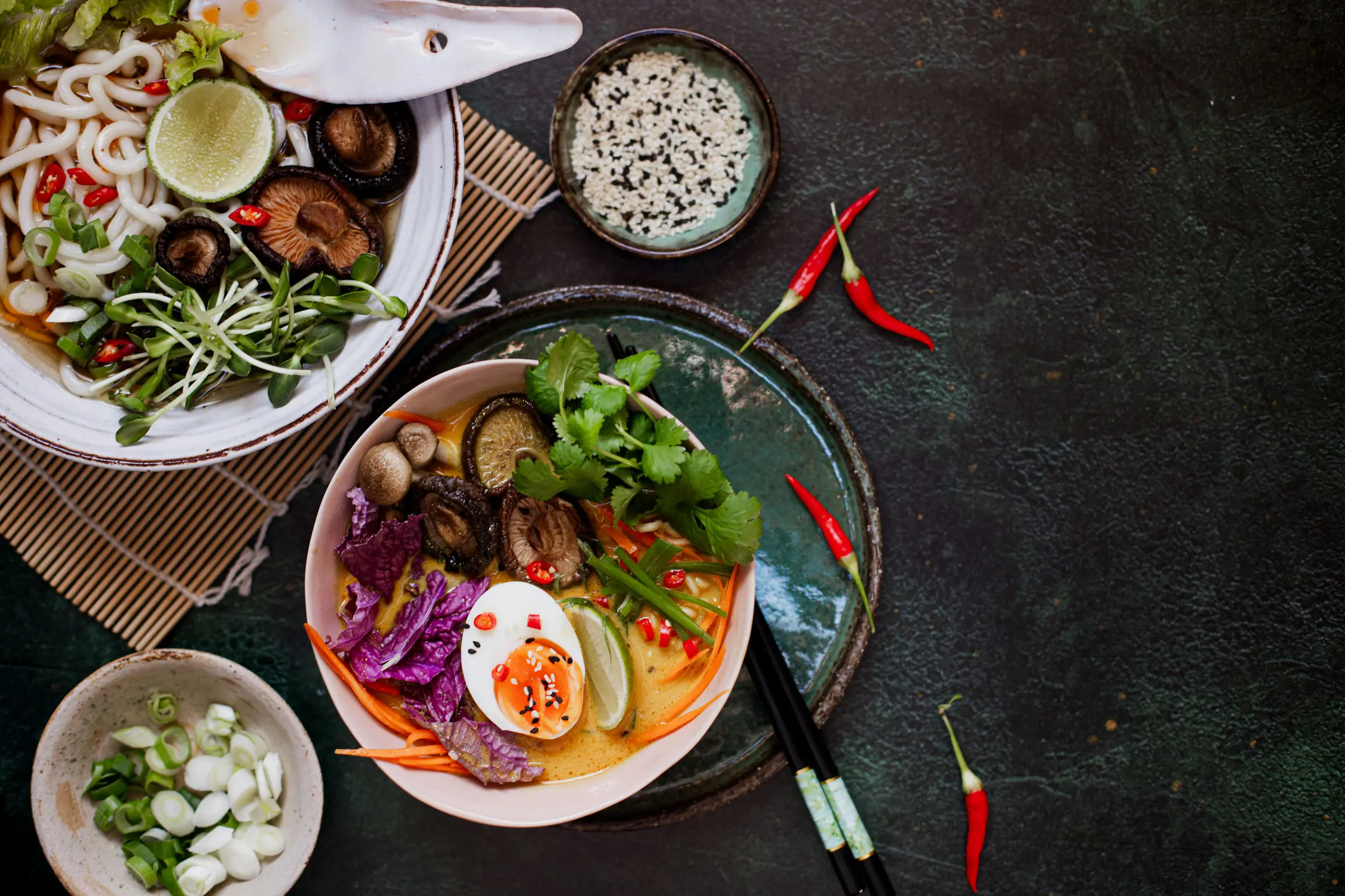 Asiatiska nudelskålar med grönsaker, lime, svamp och ägg på mörk bakgrund, dekorerade med koriander och chilipeppar.