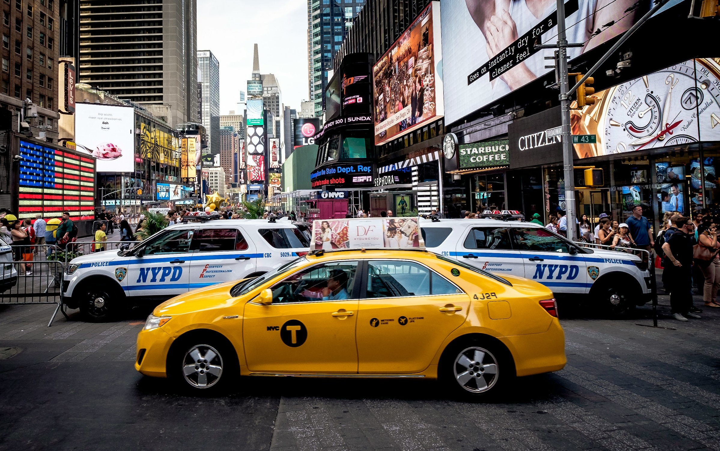 Gul taxi framför NYPD-bilar på Times Square, New York, fylld med människor och reklam.