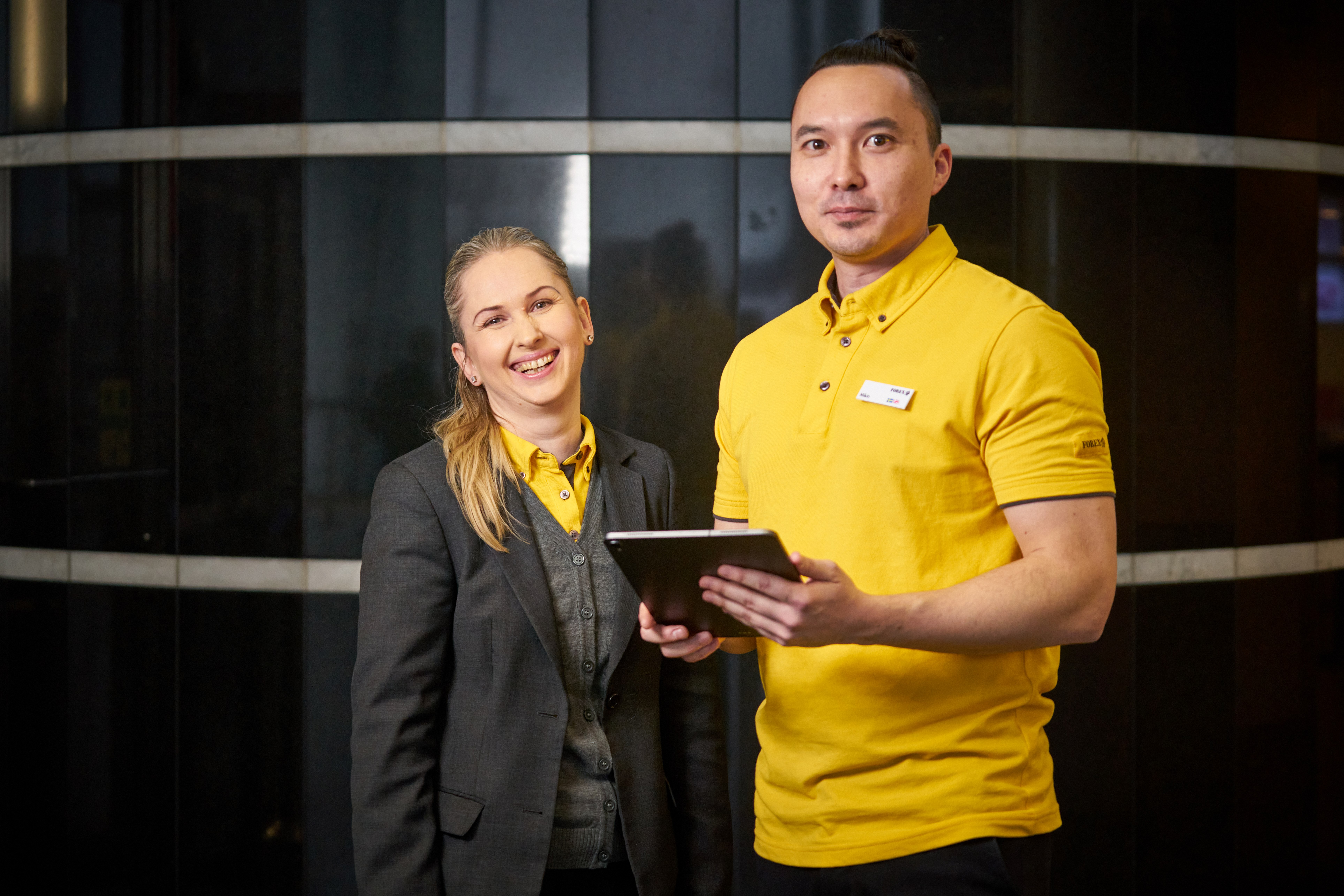 Två personer i arbetskläder håller en surfplatta framför en modern glasvägg. En person klädd i gul skjorta med namnbricka och den andra i mörk kostym med skjorta.