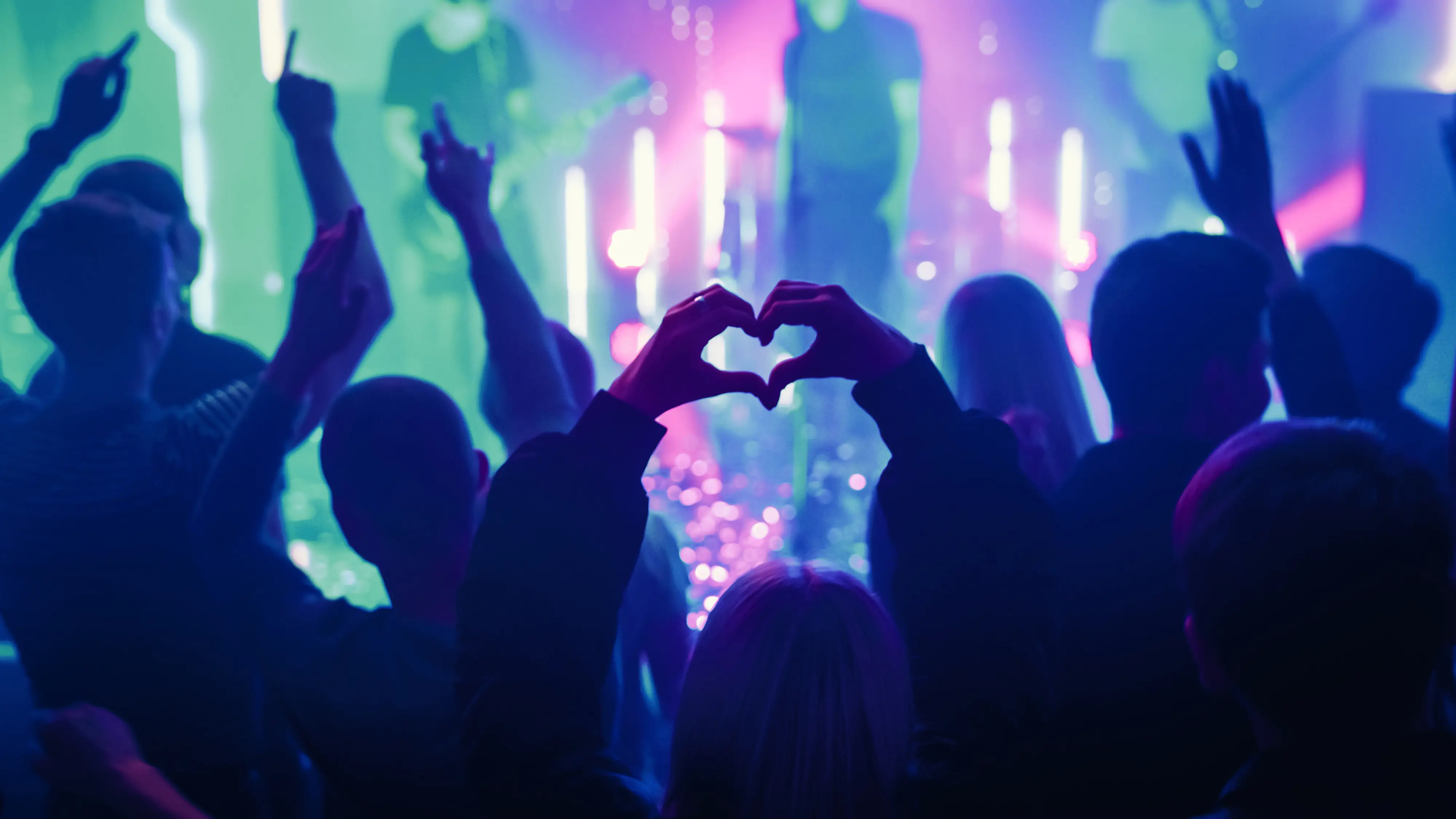 Publik njuter av en livemusikkonsert med färgglada ljus och en person som gör en hjärtform med händerna.