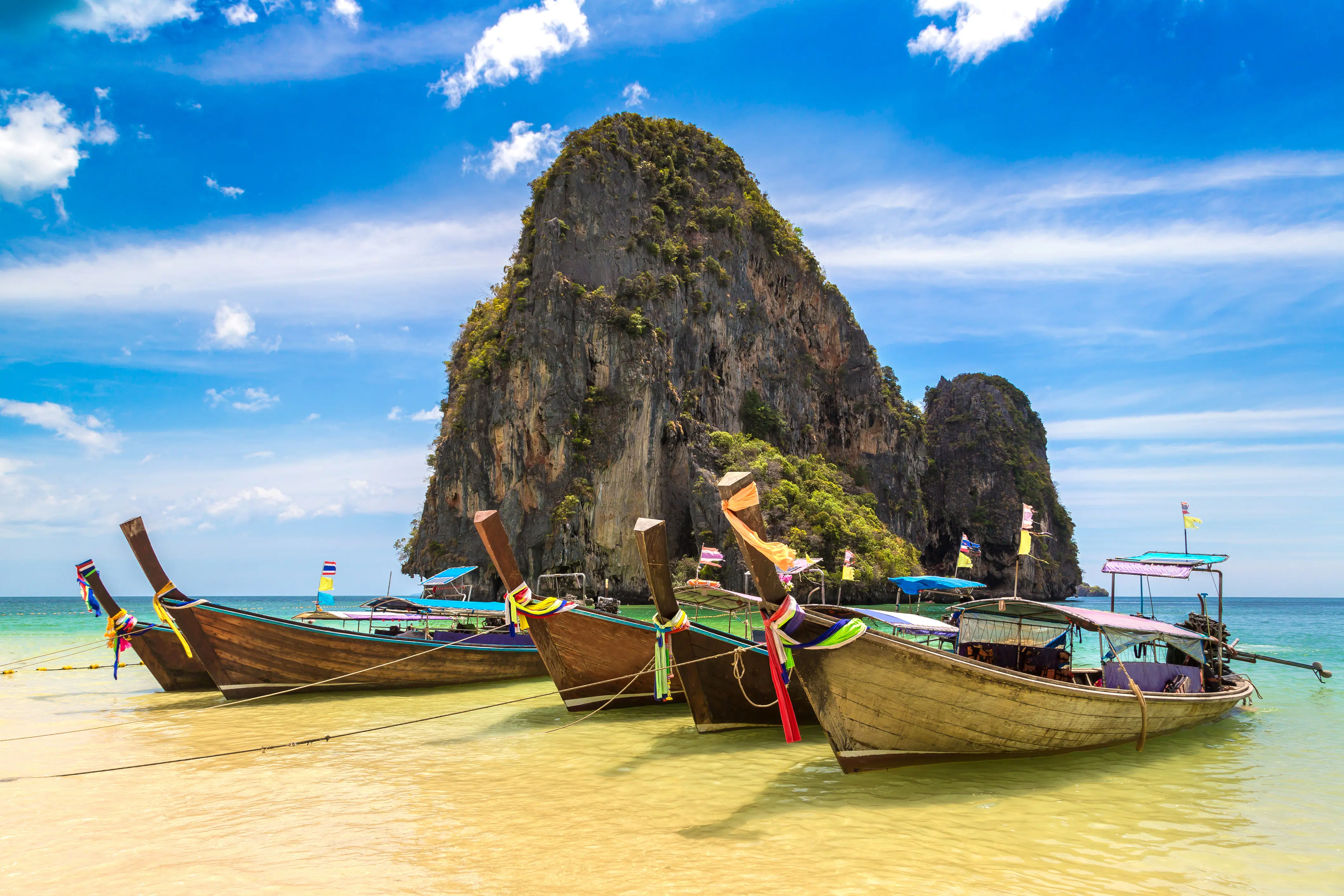 Travel to Thailand - Traditional Thai longtail boats on a sandy beach with a limestone cliff and blue sky in the background.